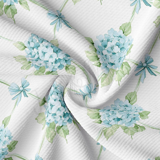 a close-up view of a fabric with a floral pattern of blue hydrangeas and green leaves. The fabric appears to be white, and the pattern is intricate and detailed.