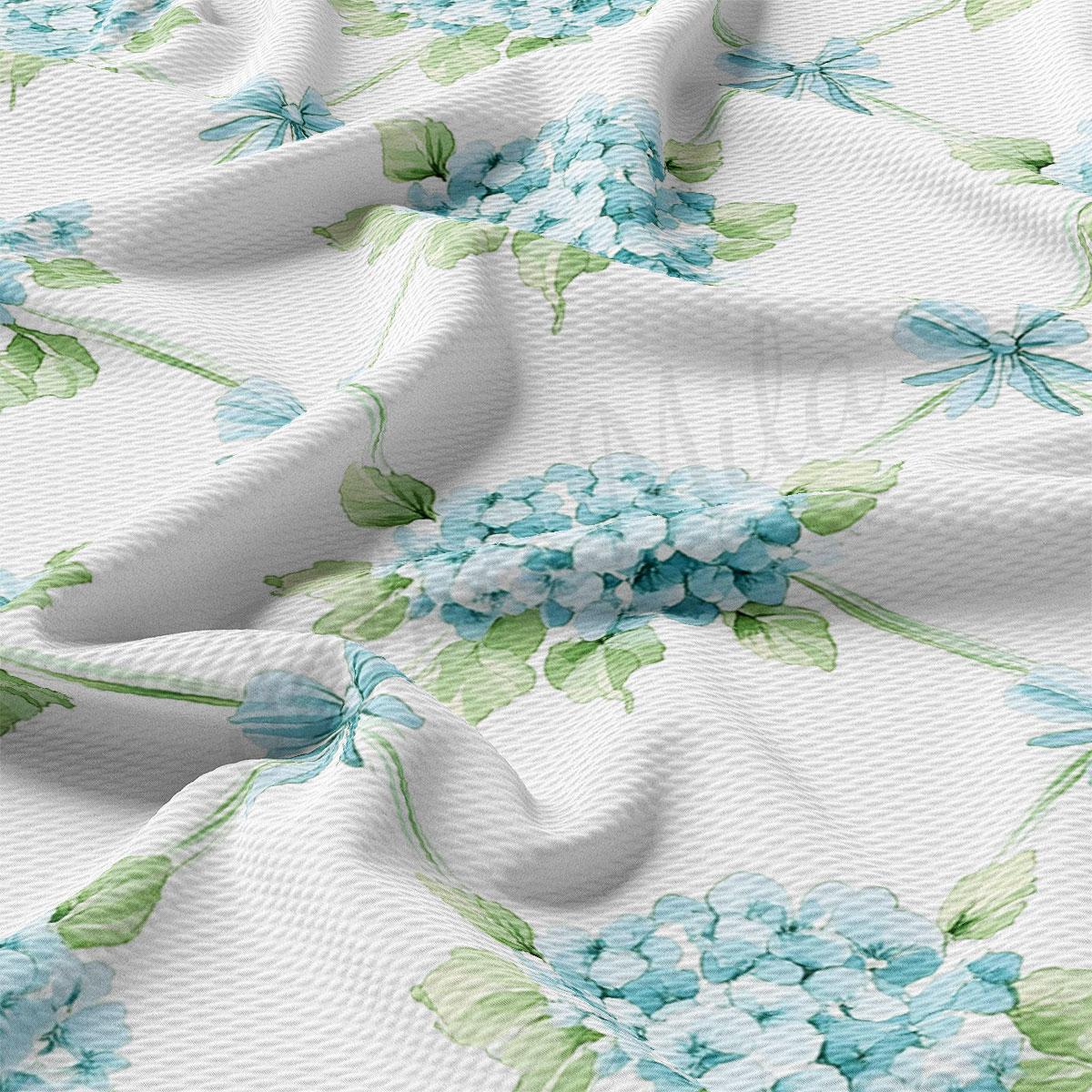 a close-up view of a fabric with a floral pattern of blue hydrangeas and green leaves. The fabric appears to be white, and the pattern is intricate and detailed, with the flowers and leaves filling the entire visible area of the fabric.
