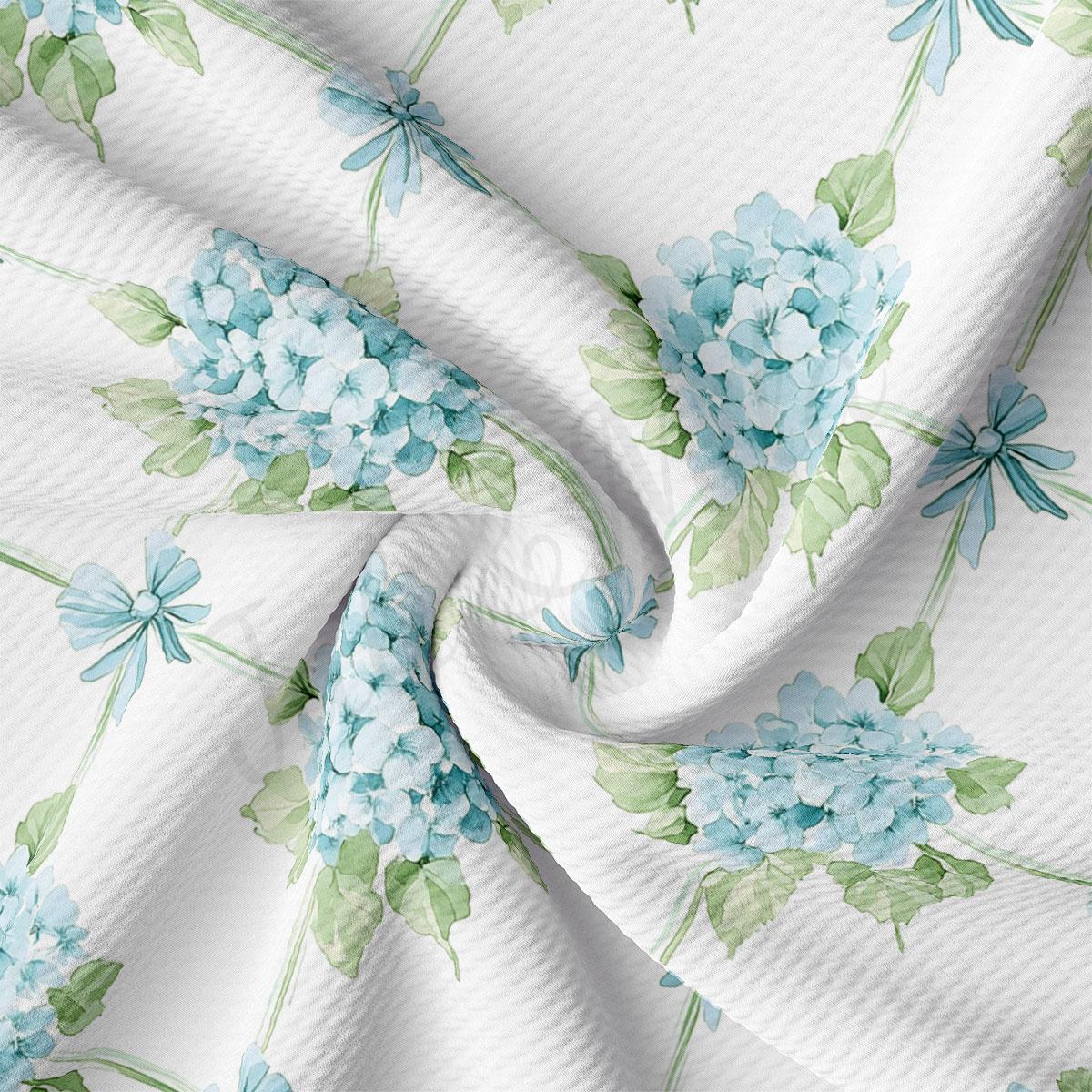 a close-up view of a fabric with a floral pattern of blue hydrangeas and green leaves. The fabric appears to be white, and the pattern is intricate and detailed.