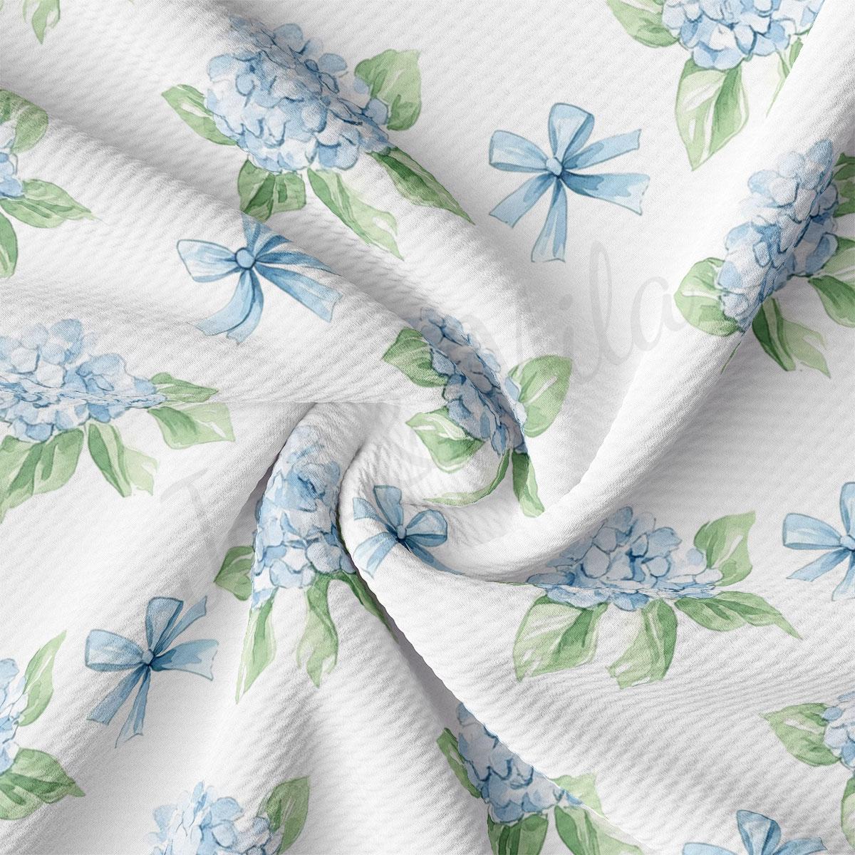 a close-up view of a fabric with a repeating pattern of blue hydrangea flowers and green leaves. The fabric appears to be white, and the pattern is intricate and detailed.