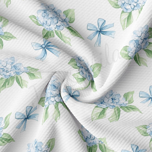 a close-up view of a fabric with a repeating pattern of blue hydrangea flowers and green leaves. The fabric appears to be white, and the pattern is intricate and detailed.