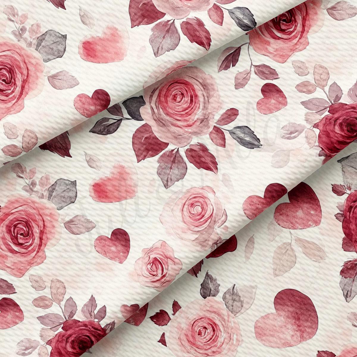 a close-up view of a fabric with a floral pattern of pink roses and hearts. The roses are the main focus, with their delicate petals and leaves filling the majority of the space. The hearts are scattered throughout the fabric, adding a touch of color and interest to the overall design. The fabric appears to be a white material, which contrasts nicely with the pink and red hues of the roses and hearts.
